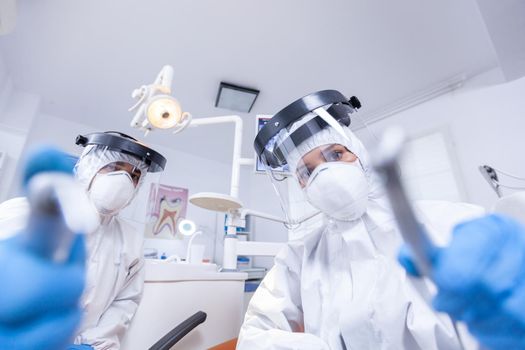 Dental team leaning over patient to work on patient gums dressed in hazmat suit. Stomatology team wearing safety equipment against coronavirus treating patient.