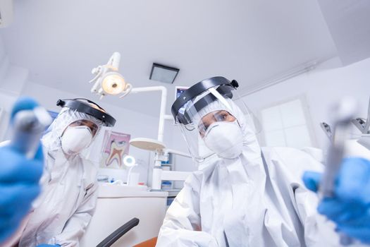 Patient pov of orthodontist and assistan doing procedure for mouth hygine in coverall. Stomatology team wearing safety equipment against coronavirus treating patient.