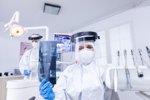 Patient pov looking at dentist showing x-ray image in dental office. Stomatology specialist wearing protective hazmat suit against coroanvirus showing radiography.