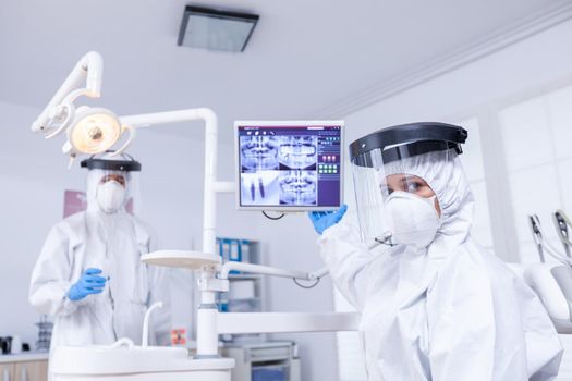 Patient pov listening explication for teeth treatment showing x-ray on monitor. Stomatology specialist wearing protective suit against infection with coronavirus pointing at radiography.