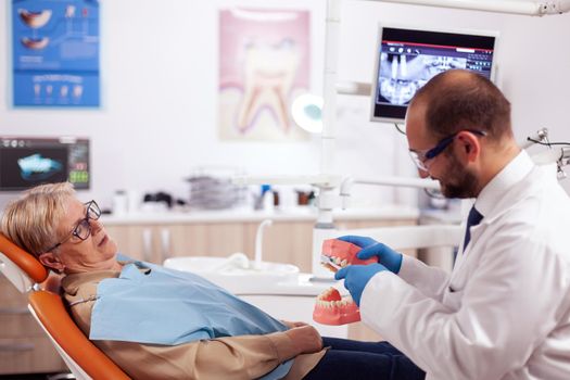 Dentist holding model of oral cavity with teeth during examination of senior patient. Medical teeth care taker discussing with senior woman about mouth hygiene.