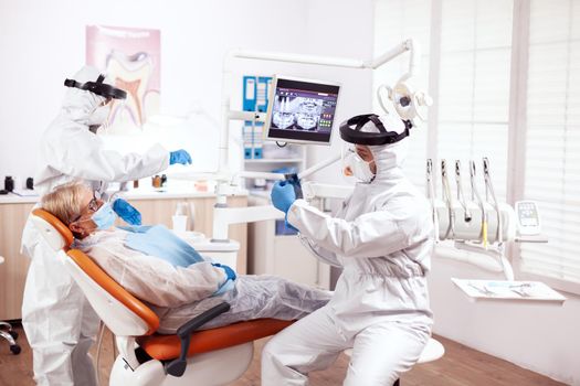 Orthodontist wearing uniform agasint coronavirus consulting senior patient. Elderly woman in protective uniform during medical examination in dental clinic.