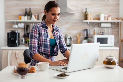 Woman smiling using laptop in kitchen in the morning with a cup of hot green tea next to her. Working from home using device with internet technology, browsing, searching on gadget in the morning.