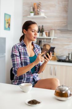 Cheerful woman doing online shopping using smartphone in kitchen and enjoing aromatic tea. Holding phone device with touchscreen using internet technology scrolling, searching on intelligent gadget.