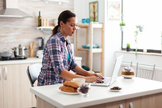 Woman typing on laptop enjoying green tea during breakfast in kitchen. Working from home using device with internet technology, browsing, searching on gadget in the morning.