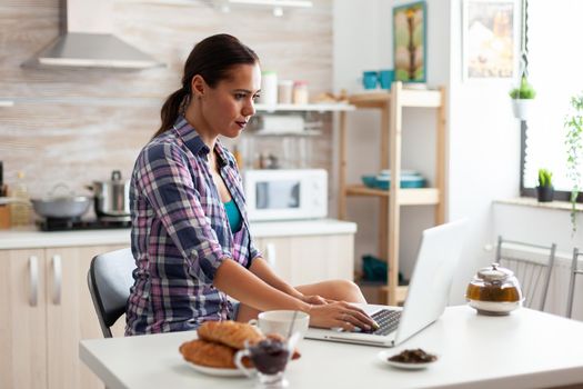 Woman working from home using laptop in kitchen during breakfast. Working from home using device with internet technology, browsing, searching on gadget in the morning.