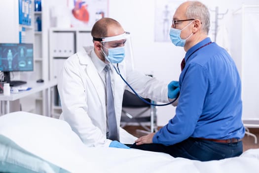 Doctor using stethoscope to listen senior man heart during examination in hospital room and wearing visor as safety precaution against coronavirus. Medical control for infections, disease.