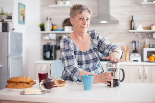 Portrait of old lady looking at camera while using french press in kitchen during breakfast. Elderly person in the morning enjoying fresh brown cafe espresso cup caffeine from vintage mug, filter relax refreshment