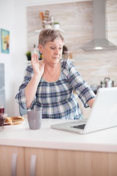 Grandmother waving during a video conference using laptop while having breakfast in kitchen. Elderly person using internet online chat technology video webcam making a video call connection camera communication conference call