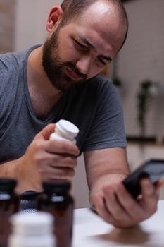 Caucasian man with headache looking at smartphone holding bottle of pills for pain remedy, treatment, medication, medicine. Adult with migraine having container with medical drugs