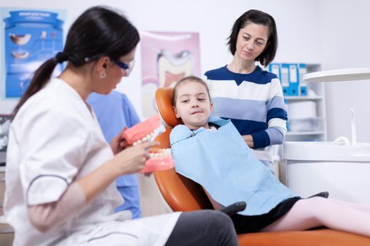 Dentist demonstrating oral hygine to little kid wearing bib and parent. Little girl and mother listening stomatolog talking about tooth hygine in dentistiry clinic holding jaw model.