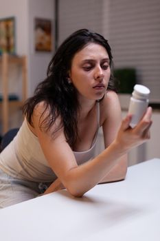 Caucasian woman with headache holding bottle of pills at home for health treatment, drugs, medication, remedy. Young adult looking at container with prescription capsules and cure