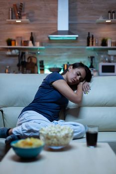 Tired lonely woman sleeping on sofa in living room while watching film. Exhausted sleepy young lady in pajamas falling asleep on sofa in front television, closing eyes at night in living room.