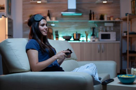 Woman gamer having fun at home sitting on sofa playing video game late at night wering eye mask on forehead. Excited determined gamer using controller joysticks keypad playstation gaming and having fun winning electronic game
