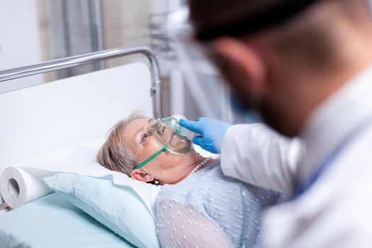 Doctor adjusting oxygen mask on senior woman while laying in hospital bed and wearing protection mask during coronavirus outbreak as safety precaution. Medicine healthcare epidemic lungs infection