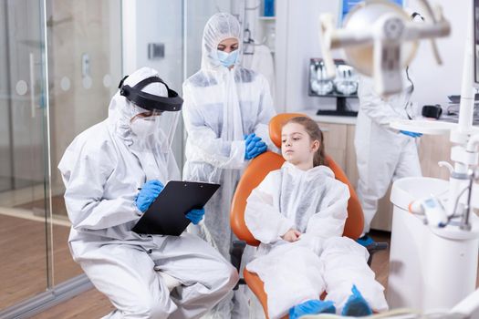 Dentist doctor in coverall taking notes on clipboard dressed in protection suit examining little girl. Stomatologist during covid19 wearing ppe suit doing teeth procedure of child sitting on chair.