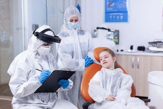 Orthodontist specialist writing treatment dressed in coverall in the course of coronavirus outbreak and kid consultation. Stomatologist during covid19 wearing ppe suit doing teeth procedure of child sitting on chair.