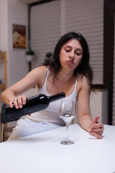 Alcoholic woman having bottle of wine and glass feeling sad at home. Lonely person drinking beverage with alcohol being depressive. Adult with addiction feeling emotional and upset