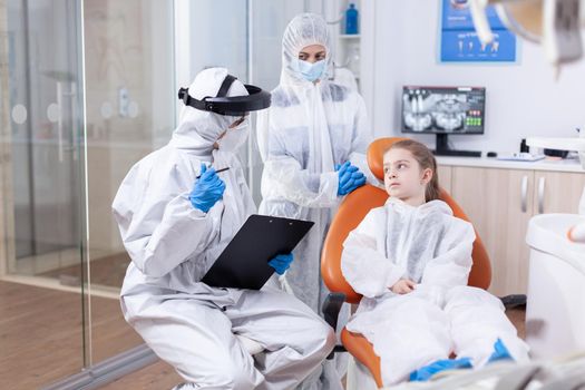 Orthodontist explaining cavity treatment to parent and little girl dressed in protection suit against coronavirus. Stomatologist during covid19 wearing ppe suit doing teeth procedure of child sitting on chair.