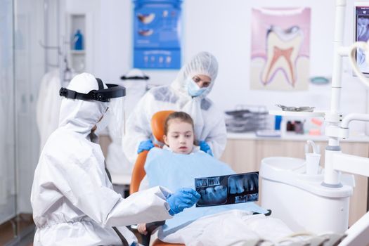Dentist dressed in ppe suit reviewing panoramic mouth radiography of little girl patient. Stomatolog in protectie suit for coroanvirus as safety precaution holding child teeth x-ray during consultation.