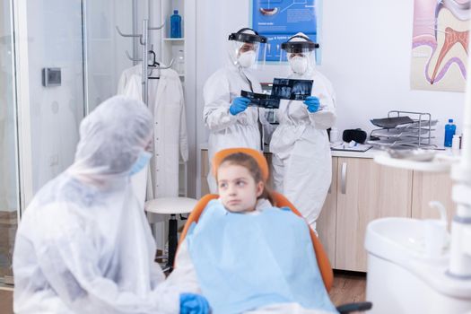 Team of dentist looking at patient dental radiography dressed in coverall in the course of coronavirus outbreak. Stomatologist during covid19 wearing ppe suit doing teeth procedure of child sitting on chair.