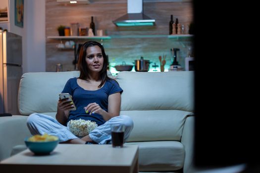 Young lady relaxing at night using phone and watching tv sitting on sofa. Lonely amused happy woman reading, writing, searching, browsing on smartphone laughing amusing using technology internet