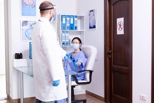 Doctor wearing protective coat and face mask against coronavirus while giving nurse patient radiography in hospital waiting room. Assistant working on reception computer.
