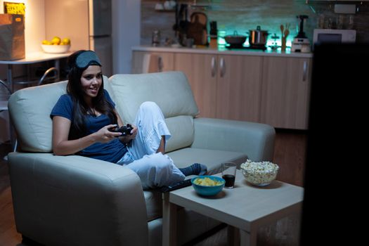 Determined woman playing video game in living room at night. Excited gamer woman sitting on couch, playing and winning video games using console and wireless controller.