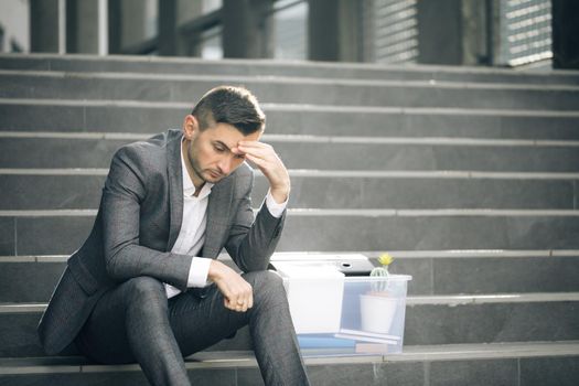 Male office worker in despair lost job. Sad businessman sitting on stairs outdoor with box of stuff as lost business. Fired man. Unemployment rate growing due pandemic.