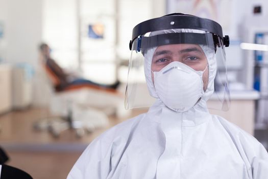 Portrait of tired doctor in dental office looking on camera wearing overall and face shield sitting on chair in waiting room clinic. Concept of new normal dentist visit in coronavirus outbreak.