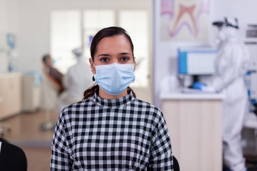 Portrait of woman in dental office looking on camera wearing face mask sitting on chair in waiting room clinic while doctor working. Concept of new normal dentist visit in coronavirus outbreak.