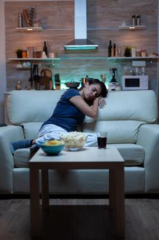 Tired woman closing eyes while watching movie at night. Tired exhausted lonely sleepy housewife in pajamas sleeping in front of television sitting on cozy couch in living room at home.