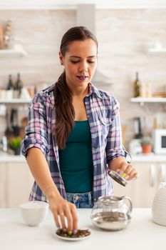 Woman using aromatic for preparation of hot tean during breakfast in home. Preparing tea in the morning, in a modern kitchen sitting near the table. Putting with hands, healthy herbal in pot.