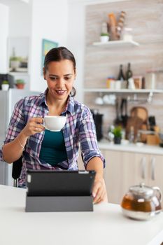 Woman smiling using tablet pc in the morning and enjoying a cup of hot green tea. Working from home using device with internet technology, typing, on gadget during breakfast.