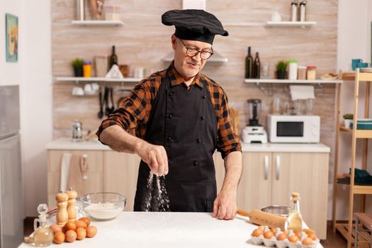Chef wearing bonete and apron while preparing ingredient for delicious recipe in home kitchen. Cook in kitchen uniform sprinkling sieving sifting ingredients by hand.