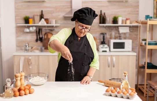 Preparation of delicious cookies in home kitchen by chef wearing apron. Happy elderly chef with uniform sprinkling, sieving sifting raw ingredients by hand.