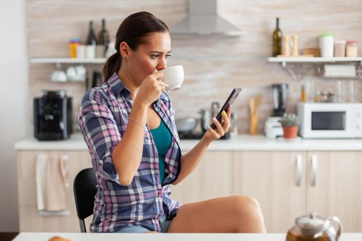 Woman drinking hot green tea in kitchen browsing on smartphone in the morning. Holding phone device with touchscreen using internet technology scrolling, searching on intelligent gadget.