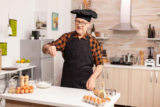 Experienced baker spreading flour in home kitchen for food preparation. Retired senior chef with bonete and apron, in kitchen uniform sprinkling sieving sifting ingredients by hand.