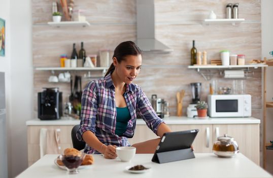 Portrait of young woman using tablet in the morning sitting at the table in the kitchen drinking tea. Working from home using device with internet technology, typing, on gadget during breakfast.