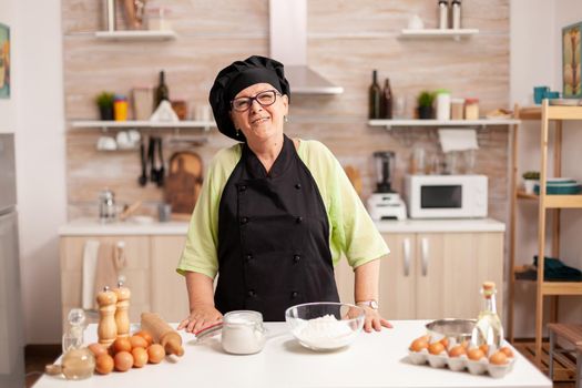 Senior woman wearing baker uniform in home kitchen smiling at camera. Retired elderly baker in kitchen uniform preparing pastry ingredients on wooden table ready to cook homemade tasty bread.