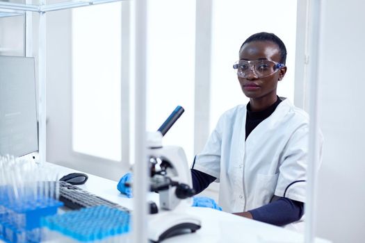 Woman of african ethnicity working in healthcare facility sitting at her workplace with protective glasses. Black healthcare scientist in biochemistry laboratory wearing sterile equipment.