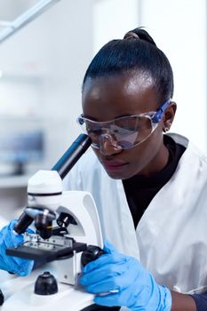 Biochemistry technician with african ethnicity using microscope for pharmacy industry with protective glasses. Black healthcare scientist in biochemistry laboratory wearing sterile equipment.