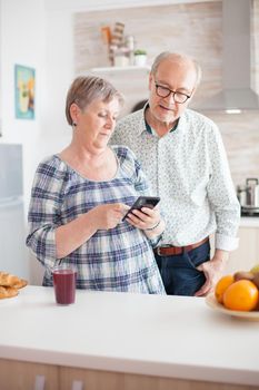 Elderly couple looking on internet and searching for information. Caucasian senior wife and husband in kitchen during breakfast using smartphone technology and internet conection. Leisure retired woman and man smiling in domicile, relaxing.