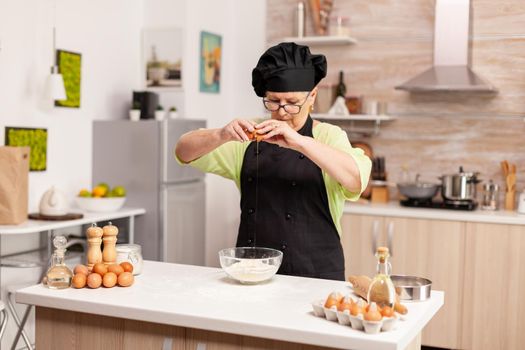 A woman prepares a dough for baking cracking eggs in home kitchen. Elderly pastry chef cracking egg on glass bowl for cake recipe in kitchen, mixing by hand, kneading ingredients prreparing homemade cake