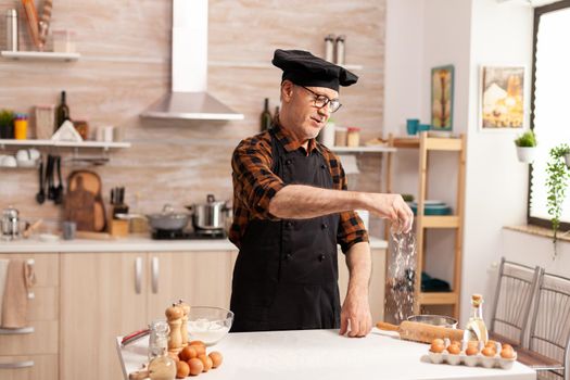 Chef in home kitchen gluten free dough for pasta, bakery or pizza. Retired senior chef with bonete and apron, in kitchen uniform sprinkling sieving sifting ingredients by hand.