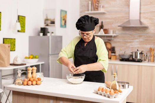 Woman breaks egg above flour making dough for bakery products. Elderly pastry chef cracking egg on glass bowl for cake recipe in kitchen, mixing by hand, kneading ingredients prreparing homemade cake