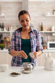 Using natural herbs in kitchen to prepare tea during breakfast. Preparing tea in the morning, in a modern kitchen sitting near the table. Putting with hands, healthy herbal in pot.