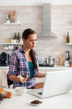 Woman looking confused at laptop screen while working from home in kitchen. Working from home using device with internet technology, browsing, searching on gadget in the morning.