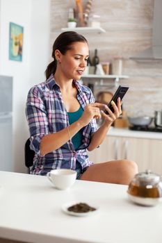 Woman scorlling on phone while having a cup of green tea in the morning. Holding phone device with touchscreen using internet technology scrolling, searching on intelligent gadget.