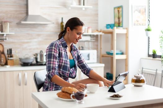 Woman browsing on tablet pc during breakfast in kitchen and holding cup of green tea. Working from home using device with internet technology, typing, on gadget during breakfast.